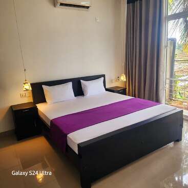A modern, minimalistic bedroom features a double bed with white linens and a purple runner, nightstands, hanging lamps, large window, curtains, and air conditioner.