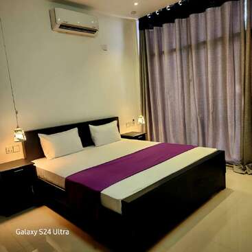 A modern bedroom with a large bed, white linens, purple runner, bedside tables, hanging lamps, air conditioner, and blackout curtains, creating a cozy, minimalist atmosphere.