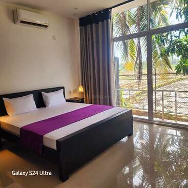 A bright, modern bedroom features a large bed with white linens and a purple runner, large windows, tropical views, curtains, air conditioner, and glossy tiled floor.