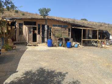 Una cabaña rústica de madera, construida a mano, se alza en una zona rural y árida. En el exterior hay barriles azules, palés y herramientas esparcidos por todas partes, que se funden con el paisaje cubierto de hierba y bañado por el sol.
