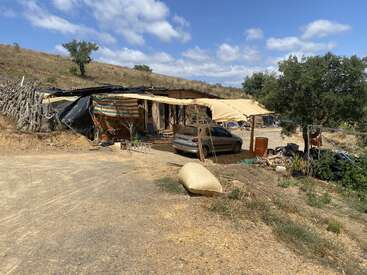 En un paisaje seco y montañoso se alza un refugio rústico construido con lonas improvisadas y postes de madera. Cerca hay un coche aparcado, rodeado de objetos diversos.