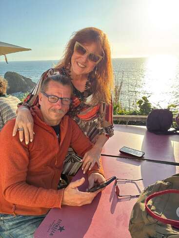 Una pareja sonriente disfruta de unas vistas soleadas al mar desde una mesa al aire libre. La mujer está de pie, mientras que el hombre está sentado con el teléfono en la mano. Con gafas de sol y un aire relajado, parecen felices.