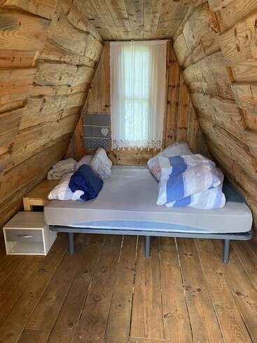Esta imagen muestra un acogedor dormitorio en una cabaña de madera con paredes inclinadas, una cama sencilla con la ropa de cama desordenada, mesitas auxiliares, suelos de madera y una ventana con cortinas.
