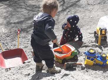 Dos niños pequeños juegan al aire libre en la tierra, rodeados de camiones de juguete, una carretilla roja y una pala. Ambos van bien abrigados y disfrutan juntos de un juego imaginativo.