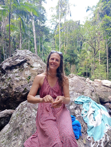 Eine Frau in einem rosa gemusterten Kleid sitzt lächelnd auf großen Felsen in einem üppigen Tropenwald. Neben ihr liegen Handtücher, was eine entspannte, natürliche Atmosphäre schafft.