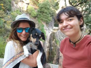 Deux femmes sourient pour un selfie en plein air, l'une tenant un chien noir et feu. Elles sont entourées de verdure luxuriante et de falaises rocheuses en arrière-plan.