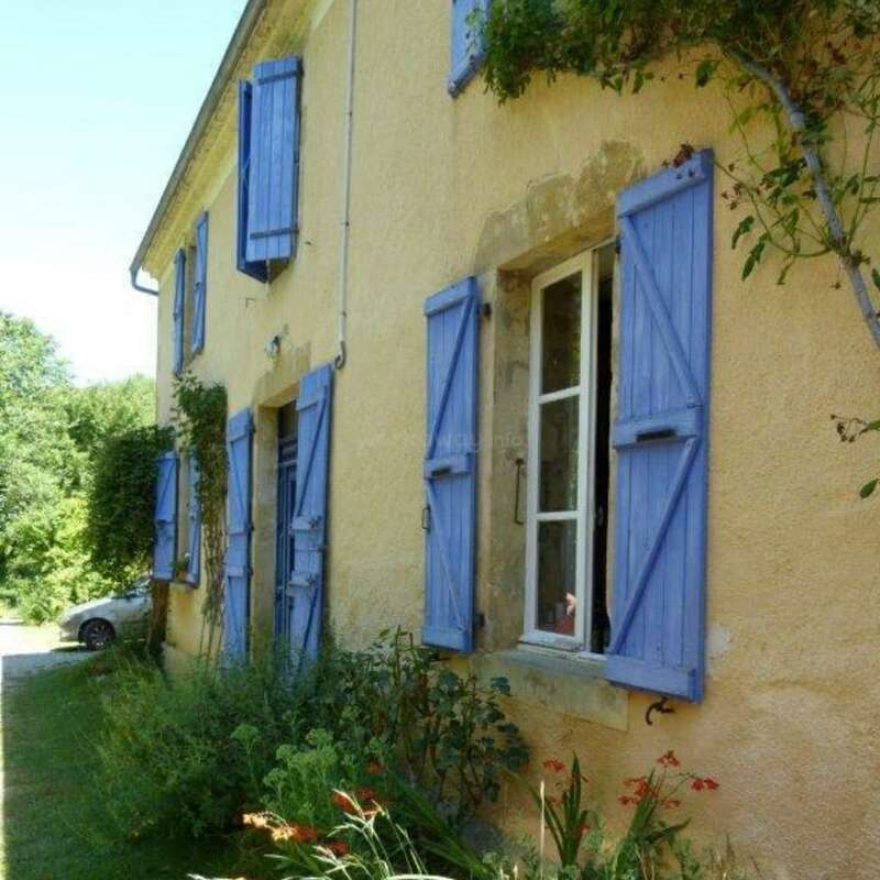 This image shows a charming yellow house with blue shutters, surrounded by lush greenery and flowers. The windows are open, letting fresh air and sunlight inside.