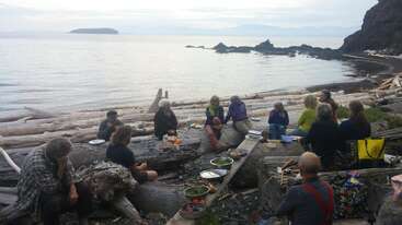 A group of people sit on driftwood logs by the seaside, enjoying a shared outdoor meal with various dishes, surrounded by a calm, scenic coastline.