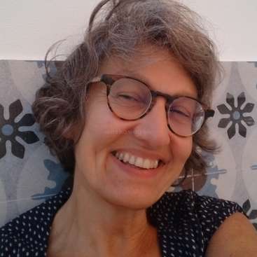 The image shows a woman with short gray hair, smiling at the camera, wearing glasses and a dark blue top with white dots, against a patterned wall.