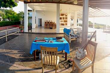 Una terraza al aire libre con una mesa cubierta con un mantel azul sobre la que se aprecian equipos musicales, sillas de madera y un instrumento de cuerda tradicional, con un edificio moderno, espacioso y bañado por la luz del sol como fondo.