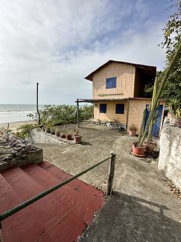 Una rústica casa de playa de madera de dos plantas, con ventanas azules, se alza junto al mar. Macetas con plantas y cactus decoran el patio de hormigón bajo un cielo nublado.