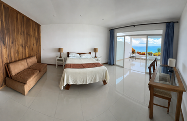 Esta amplia y moderna habitación cuenta con una pared decorativa de madera, una cama, un sofá marrón, un escritorio, amplios ventanales y unas preciosas vistas al mar con una zona de descanso al aire libre.