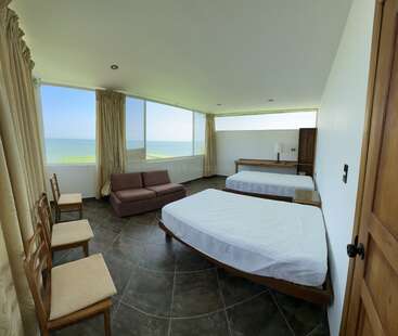 Esta amplia y moderna habitación de hotel cuenta con dos camas, un sofá, tres sillas y grandes ventanales con cortinas, lo que ofrece unas impresionantes vistas al mar y mucha luz natural.