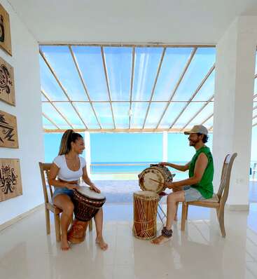 Dos personas están sentadas una frente a otra, sonriendo y tocando la batería en una sala luminosa y diáfana con vistas al mar, rodeadas de una decoración artística en las paredes y de la luz natural del sol.