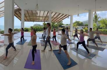 Un grupo de personas practica yoga juntas sobre colchonetas de colores en un pabellón abierto y espacioso, con unas vistas panorámicas de la vegetación, las montañas y el mar al fondo.