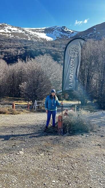Una mujer vestida de invierno junto a una bandera de "La Montañita Equipo Outdoor", con montañas nevadas, árboles desnudos y un cielo azul despejado de fondo.