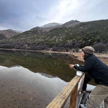Una persona, de pie sobre una plataforma de madera, contempla pensativa un lago en calma. Las montañas y los bosques se reflejan en el agua bajo un cielo nublado.