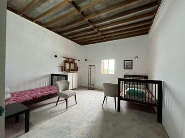 Cette chambre comprend deux lits simples, deux chaises, une petite table, des étagères et une décoration épurée. Le plafond brut et les murs blancs sobres créent une ambiance rustique.