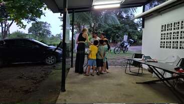 A group of children and an adult gather outside under a roof, near a white building with tables, chairs, a parked car, and a motorcycle.