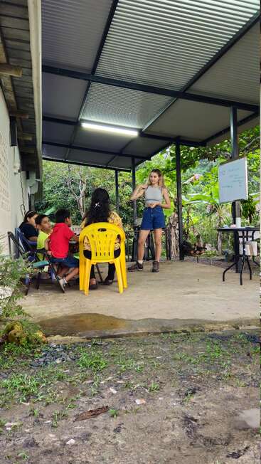 A young woman stands teaching a group of children seated outside on a covered patio, surrounded by nature, with a whiteboard and green trees nearby.