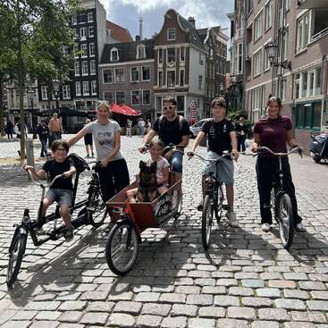 Une famille de six personnes, composée de deux adultes, trois enfants et un chien, fait du vélo ensemble dans une rue pavée d'une charmante ville européenne.