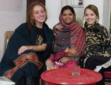 Three women sit together smiling, dressed warmly in colorful, patterned shawls and sweaters. They appear relaxed, sitting indoors beside a red table with a glass.
