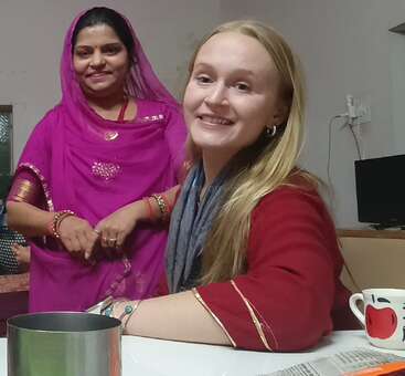 Two women smiling indoors, one in a bright pink traditional outfit and the other in red, sitting at a table with a cup and a newspaper.