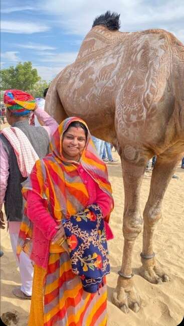 The image depicts a woman in traditional Indian attire standing beside a large statue of an animal, possibly a camel, on a sandy surface.