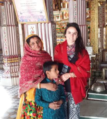 The image depicts two women and a child in a room with striped wallpaper, featuring a woman in a red shawl, a woman in a pink shawl, and a child in a blue shirt.