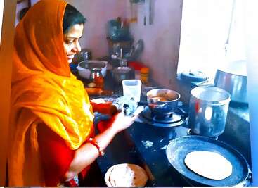 The image depicts a person wearing an orange head covering and red clothing, leaning over a black stove with a pot in their hand.
