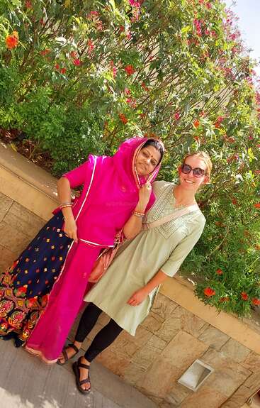 Two women, one in vibrant traditional Indian attire and the other in a light-colored dress with sunglasses, smiling together outdoors against a backdrop of flowering bushes.