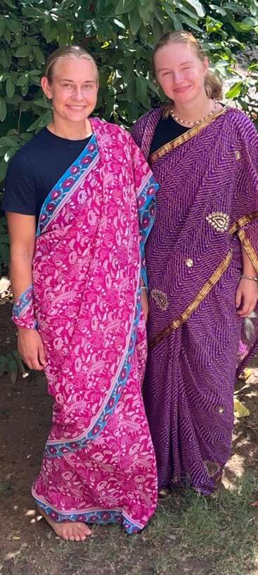 Zwei Frauen stehen lächelnd im Freien, gekleidet in farbenfrohe traditionelle indische Saris – die eine in Rosa und Blau, die andere in Lila und Gold – unter üppigem grünem Laub.