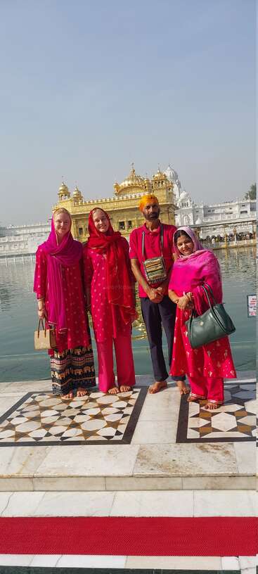 Vier Menschen in farbenfroher traditioneller Kleidung stehen barfuß vor dem Goldenen Tempel in Amritsar, hinter ihnen das ruhige Wasser und der klare Himmel.