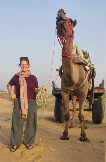 Una mujer sonriente está de pie en el desierto, sujetando la rienda de un camello. El camello lleva un arnés rosa y se encuentra junto a una carreta, bajo un cielo despejado.