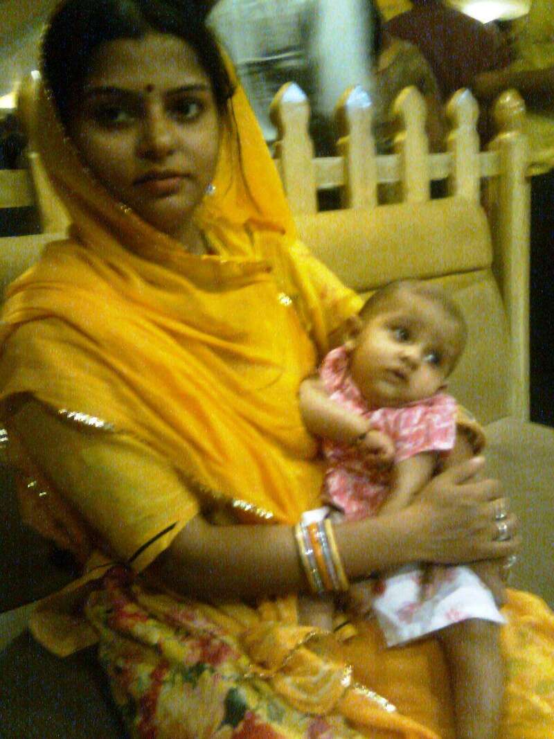 The image depicts a woman in a yellow headscarf holding a baby in a pink outfit, with a wooden fence and other objects in the background.