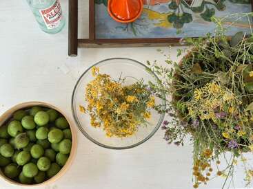 Sobre una mesa blanca: un cuenco de frutas verdes, un cuenco de cristal con flores amarillas, flores silvestres secas, una botella, una naranja exprimida y una bandeja de colores.