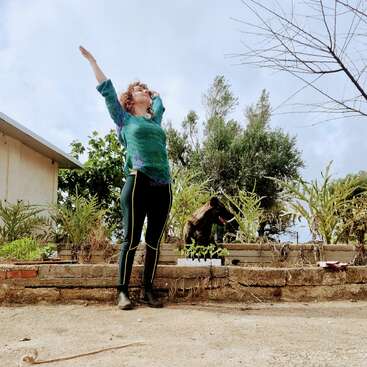 Uma mulher está ao ar livre em uma saliência de pedra, com os braços levantados alegremente em direção ao céu. Ela está cercada por plantas, árvores, uma casa e um cachorro.