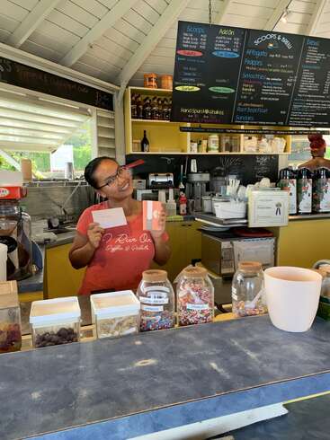 Una mujer sonriente detrás del mostrador de una heladería con tarjetas de regalo en la mano. En unos tarros se exhiben coloridos polvorones y coberturas. El menú cuelga del techo.