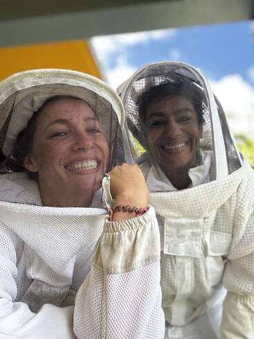 Dos mujeres sonríen alegremente, vestidas con trajes de apicultura de malla blanca y velos protectores. Parecen felices, al aire libre, posiblemente trabajando juntas en actividades apícolas.