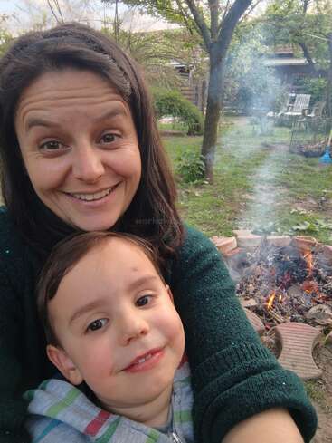 A woman and young child smile for a selfie outdoors beside a campfire. The background features trees, grass, a rake, and a cozy garden setting.