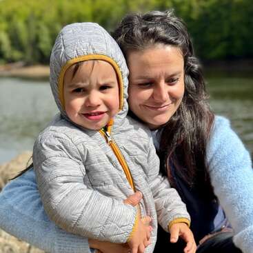 La imagen muestra a una mujer y un niño posando para una foto a orillas de un lago, con el niño vestido con una chaqueta gris y la mujer con un jersey azul.