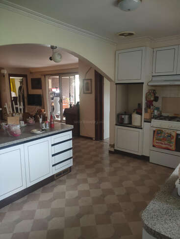The image depicts a kitchen with white cabinets and countertops, featuring a stove, sink, and refrigerator, with a doorway leading to a living room and patio area in the background.