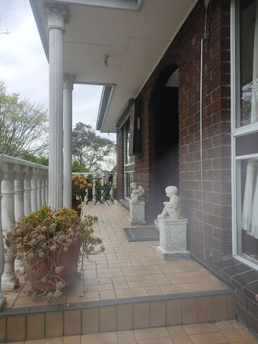 The image depicts the exterior of a house with a covered porch, featuring white columns, a tiled floor, and a brick facade, surrounded by trees and a cloudy sky.