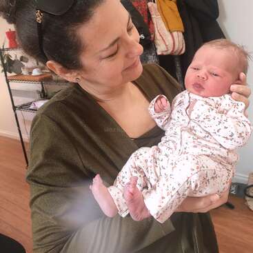 La imagen muestra a una mujer acunando a un bebé en brazos, con el pelo oscuro de la mujer recogido en un moño y el bebé con un body blanco de flores.