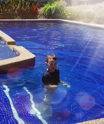 The image depicts a young boy standing in a pool, wearing a dark blue shirt, with a poolside garden in the background and a sunny day atmosphere.