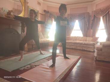 Two people are standing on yoga mats in a living room, with their arms outstretched, wearing dark clothing, and facing the camera.
