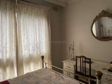 A cozy bedroom with light floral curtains, a white bed frame, ornate mirror, classic desk, lamp, chair, and soft natural light filtering through the window.