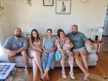 Five adults and a baby sit closely together on a light-colored couch in a cozy living room, smiling warmly, with art and plants in the background.