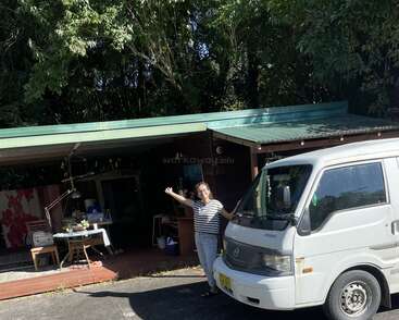 Une femme se tient à côté d'une camionnette blanche devant une maison rustique au toit vert, entourée d'arbres luxuriants, profitant d'une journée ensoleillée en plein air.