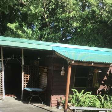 Uma pequena casa de madeira com um telhado de metal verde é cercada por uma vegetação exuberante. Há uma mesa do lado de fora, painéis de treliça e samambaias perto da entrada.